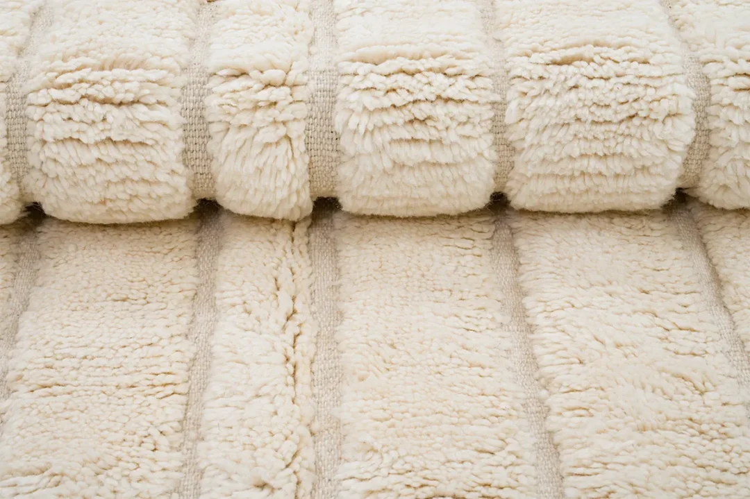 A close-up view of a cream-colored, plush, textured blanket or rug with thick, soft ridges and a fluffy appearance.