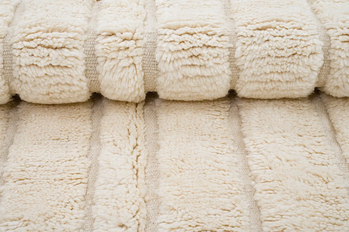 A close-up view of a cream-colored, plush, textured blanket or rug with thick, soft ridges and a fluffy appearance.
