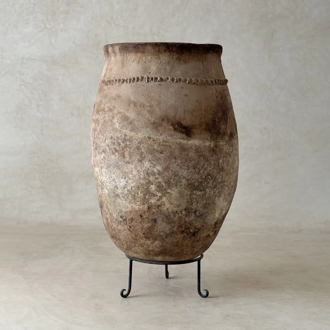 Large, weathered clay vessel with a rounded body and narrow neck, featuring a decorative ridge, displayed on a minimalist black metal stand against a neutral background.
