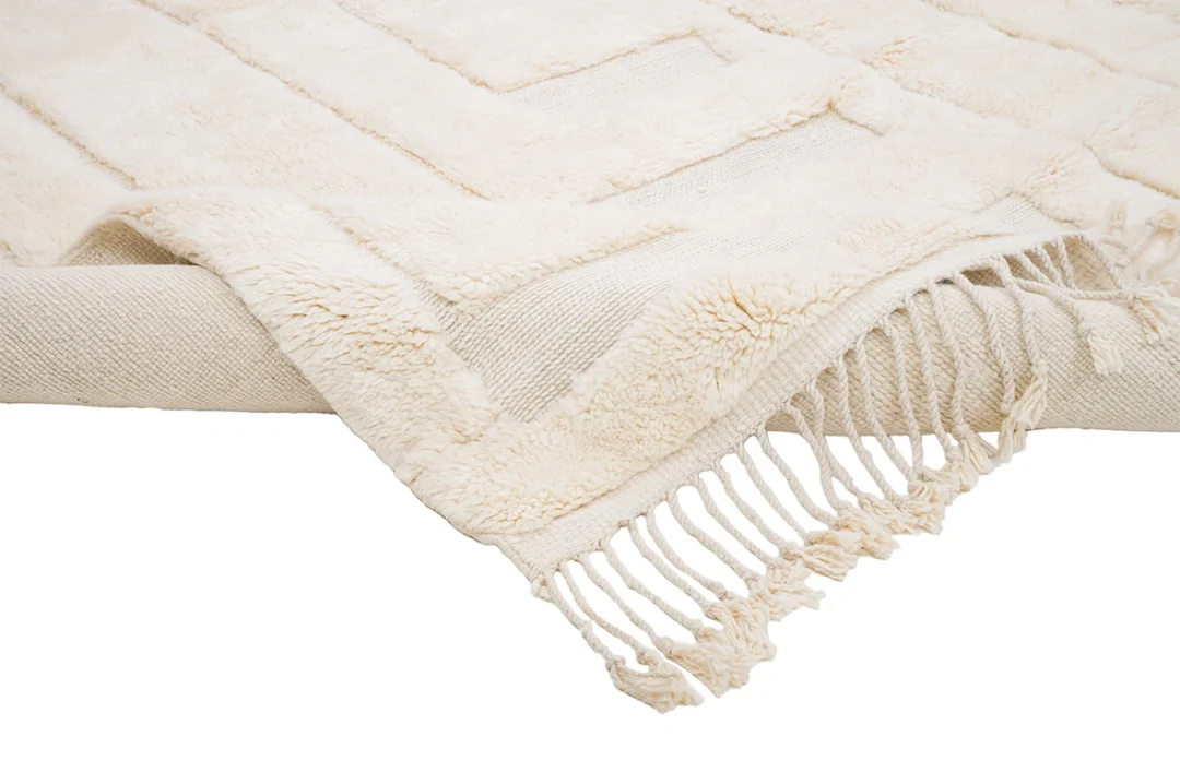 A cream-colored textured throw rug with a geometric pattern and fringe detail draped over the edge of a surface.