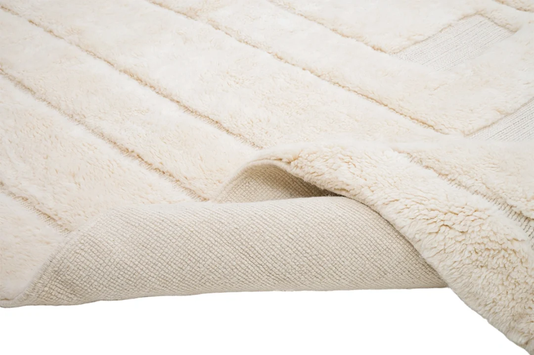 A close-up of a cream-colored, textured rug with one corner flipped over, showing the woven underside and soft, plush surface.
