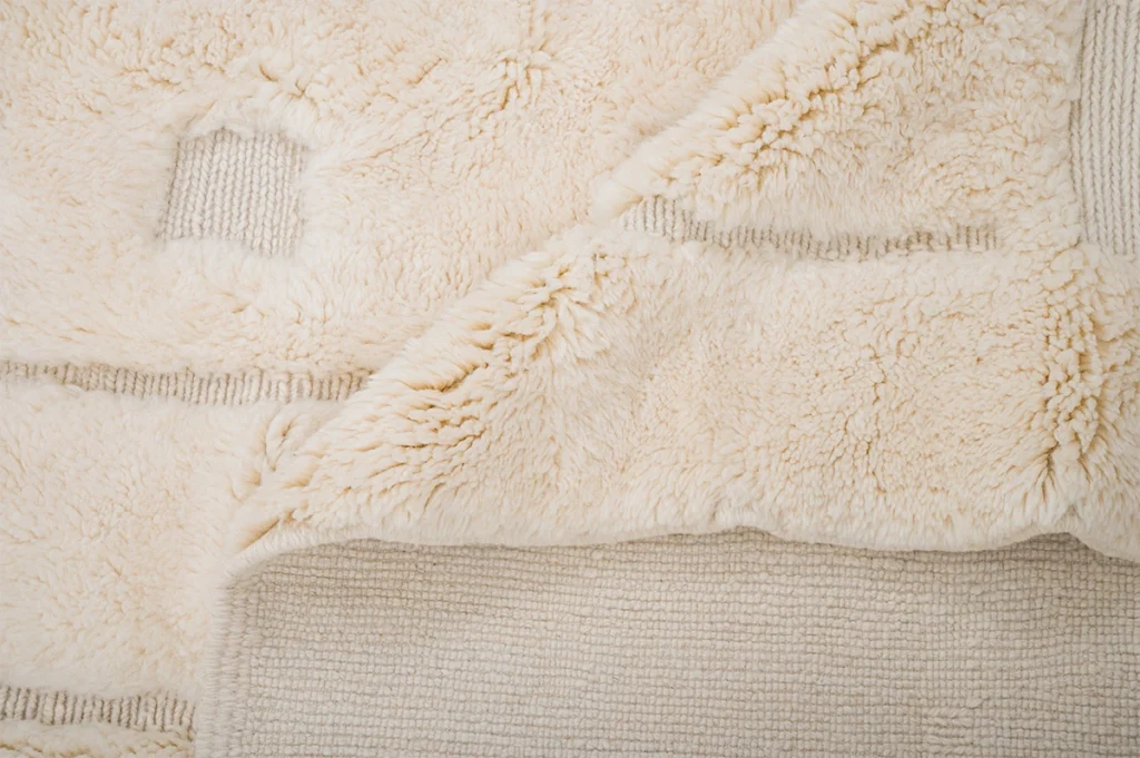 A close-up of a cream-colored, textured rug with a soft, fluffy surface and a corner folded to reveal the woven underside.
