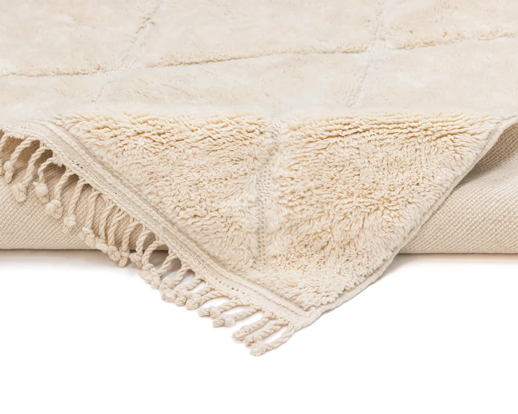 Close-up of a beige rug with a textured pattern, fringed edges, and a soft, plush surface corner folded over.