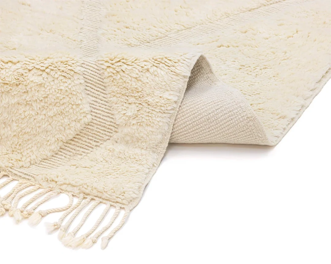 A cream-colored textured rug with tasseled edges is shown partially folded to display both the front and the woven back.