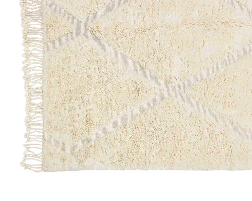 A close-up of a cream-colored textured rug with a subtle geometric pattern and fringed edges on one side.