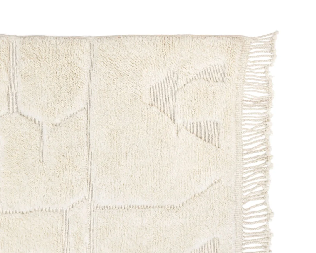 A close-up of a cream-colored textured rug with subtle geometric patterns and fringe along the right edge.
