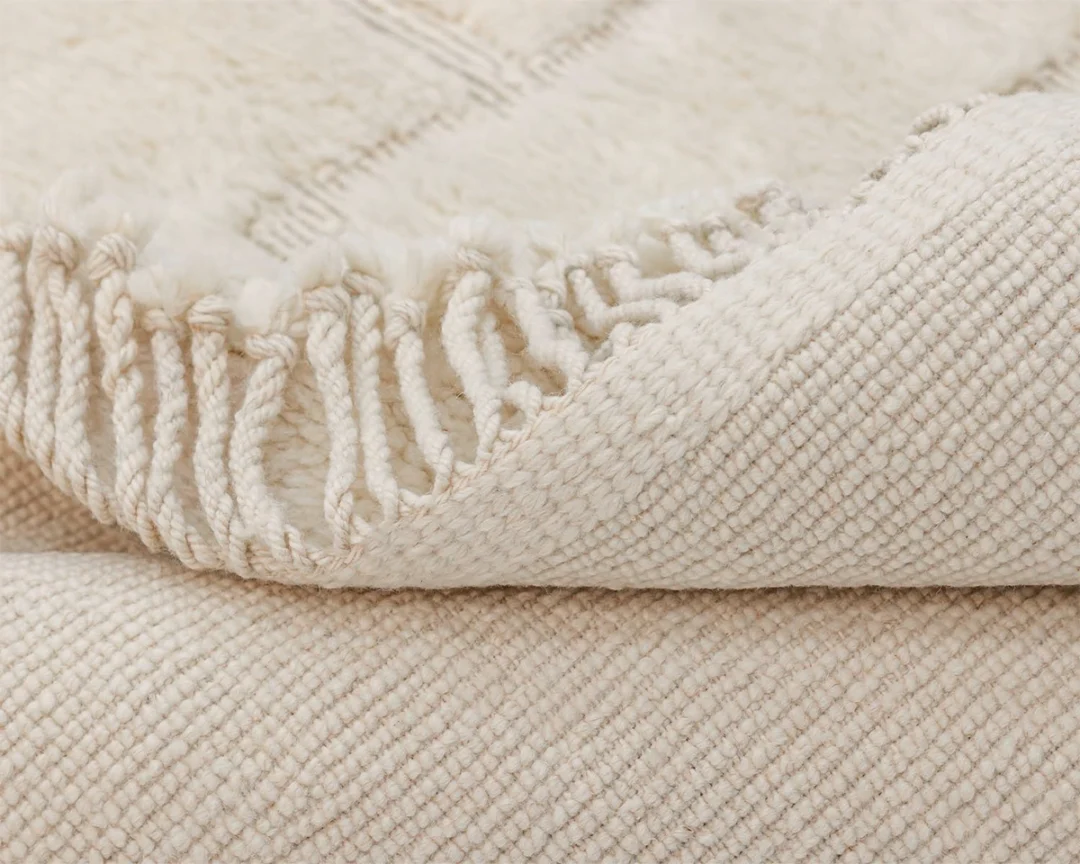 Close-up of a cream-colored woven blanket with a fringe edge, showing its textured surface and soft, cozy appearance.