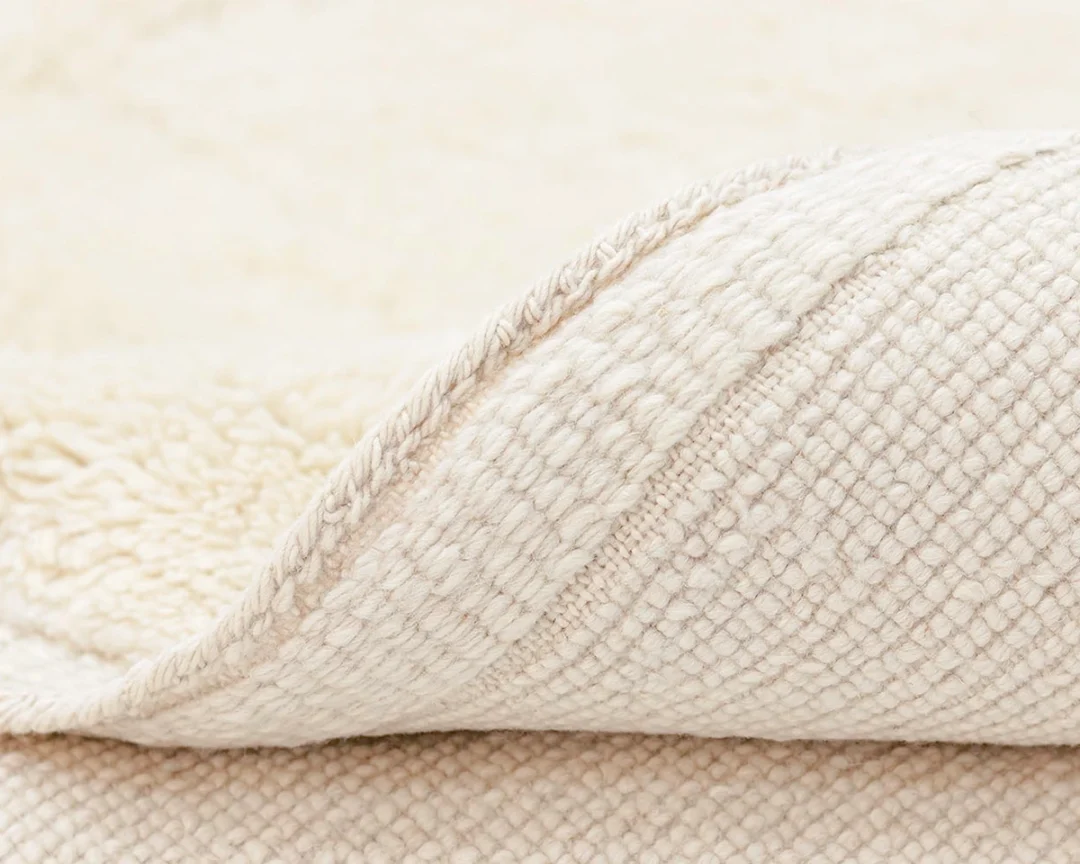 A close-up of a cream-colored textured rug shows a soft, woven surface and a plush inner lining, with the edge slightly folded over.