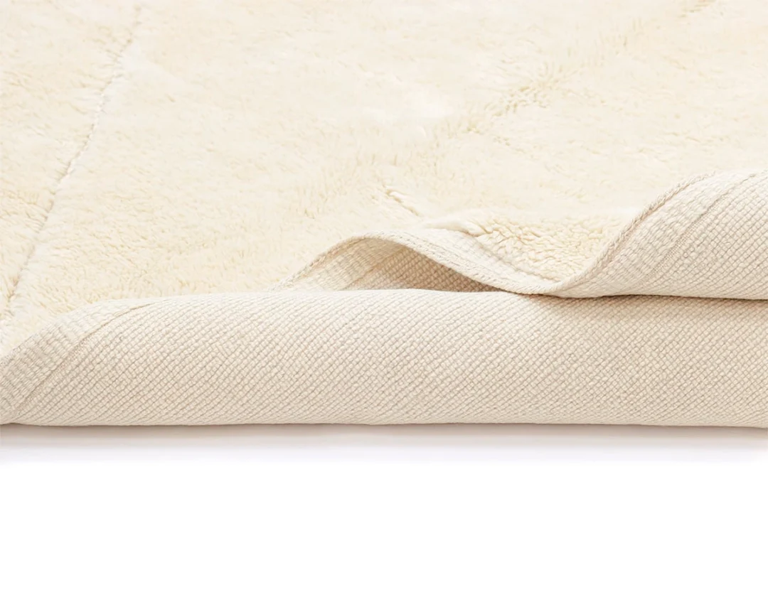 A close-up view of a cream-colored, textured rug with one corner folded over to show both the soft top surface and the woven underside.