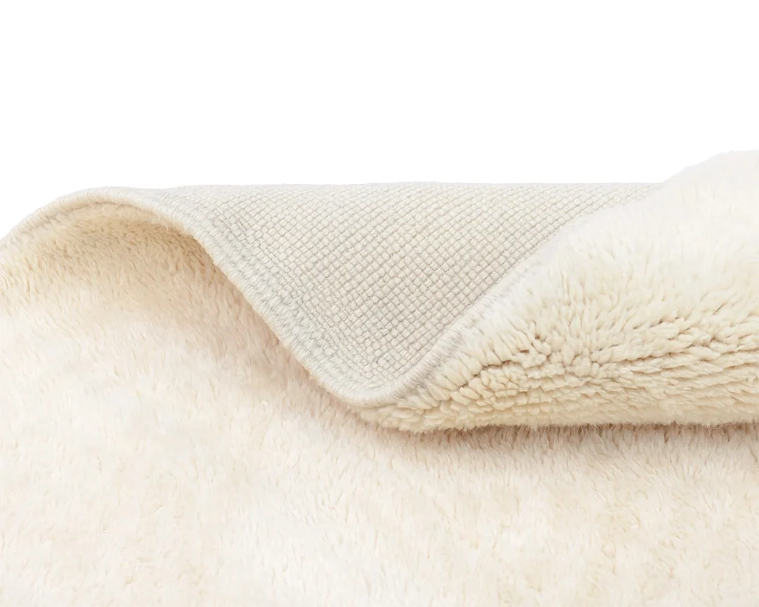 A close-up of a cream-colored rug folded to show its soft, fluffy texture on one side and a textured, woven backing on the other.