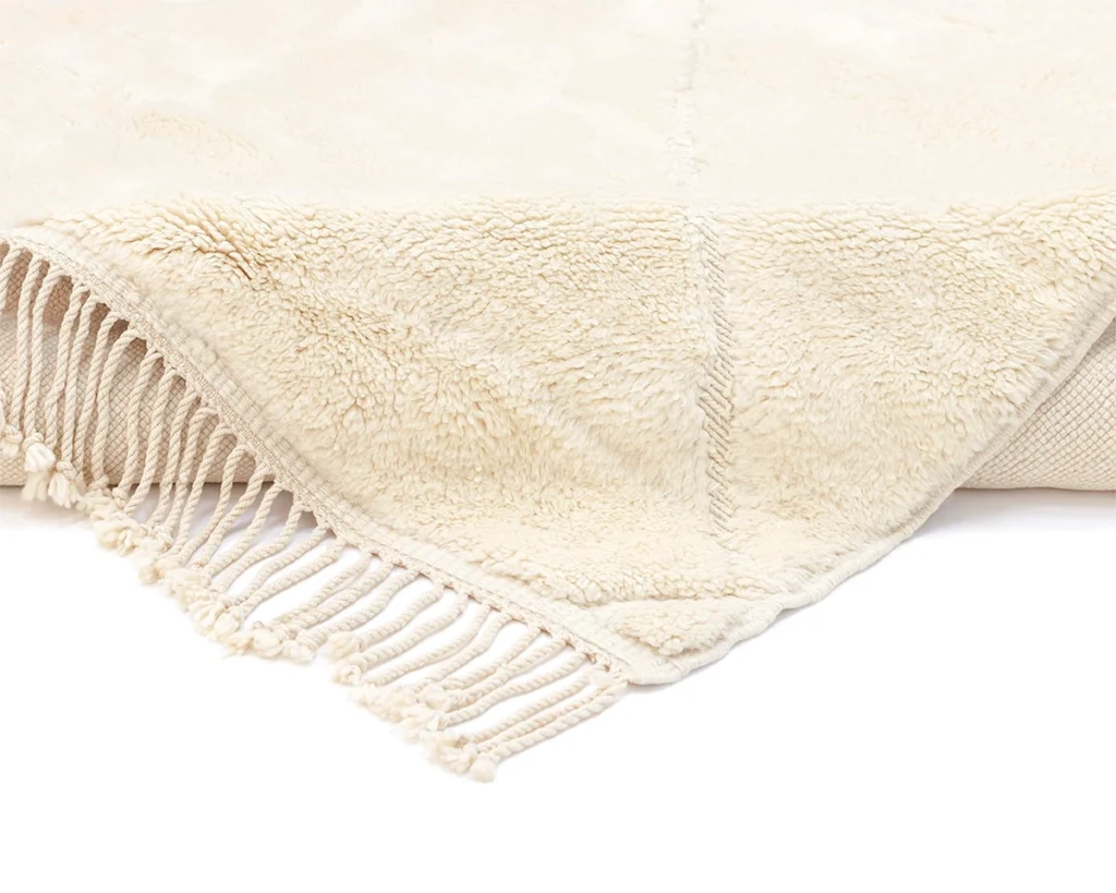 A close-up of a beige, fringed throw blanket with a soft, textured surface and stitched detailing.