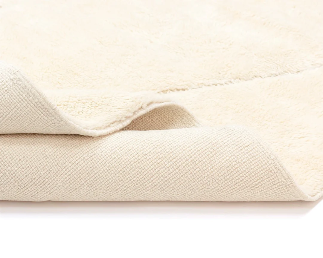 A close-up view of a cream-colored, textured rug partially folded to show both the soft, plush top and the woven underside.