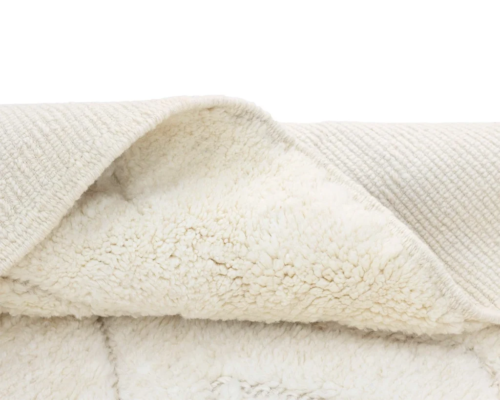 Close-up of a cream-colored rug with a textured top layer and a thick, fluffy underside, partially folded over to show both surfaces.