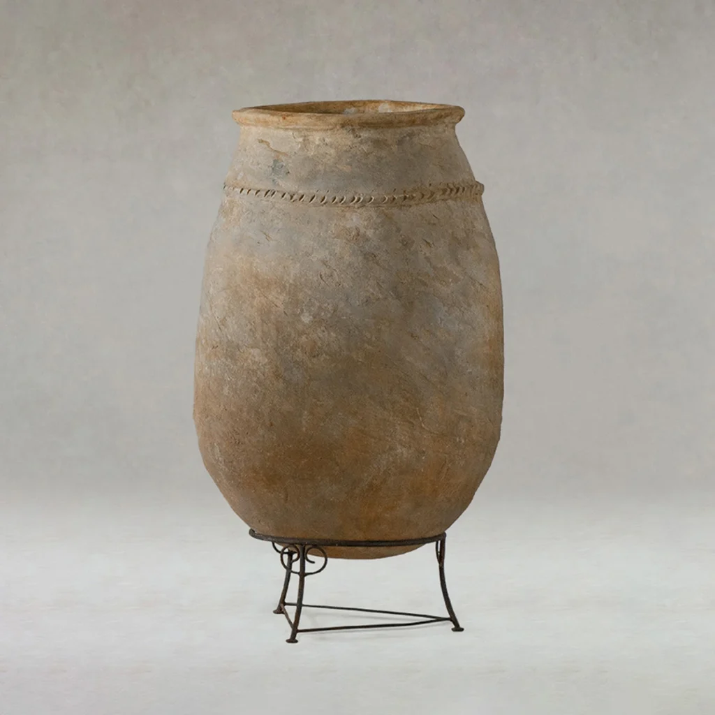 Large, weathered clay storage jar with a rope-like decorative band near the top, displayed on a simple metal stand against a neutral background.