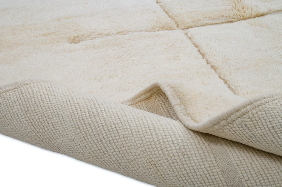 A close-up of an off-white, plush area rug with a textured underside, showing one corner slightly folded over.