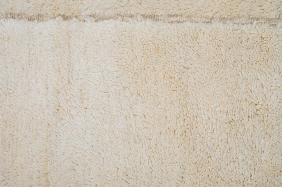 Close-up view of a beige, textured, shaggy rug with visible fibers and a horizontal seam near the top.