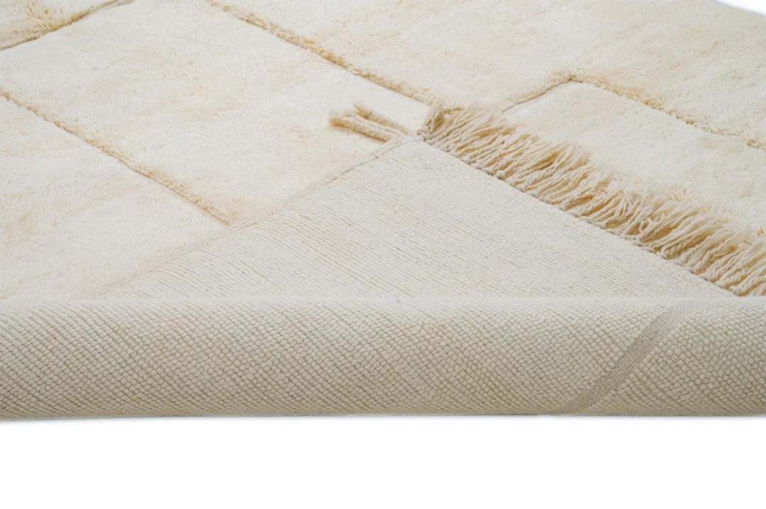 Close-up of a beige textured rug with a folded corner, showing fringed edges and various weaves and patterns.