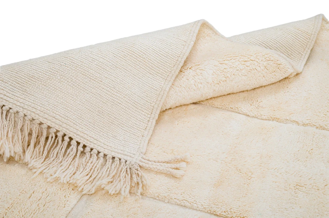 A beige rug with a textured surface, tasseled fringe on one end, and a visible soft underside, shown partially folded on a white background.