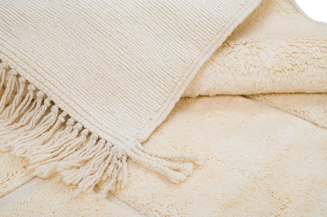 Close-up of a cream-colored woven blanket with fringe detail, layered over a soft, plush beige blanket.