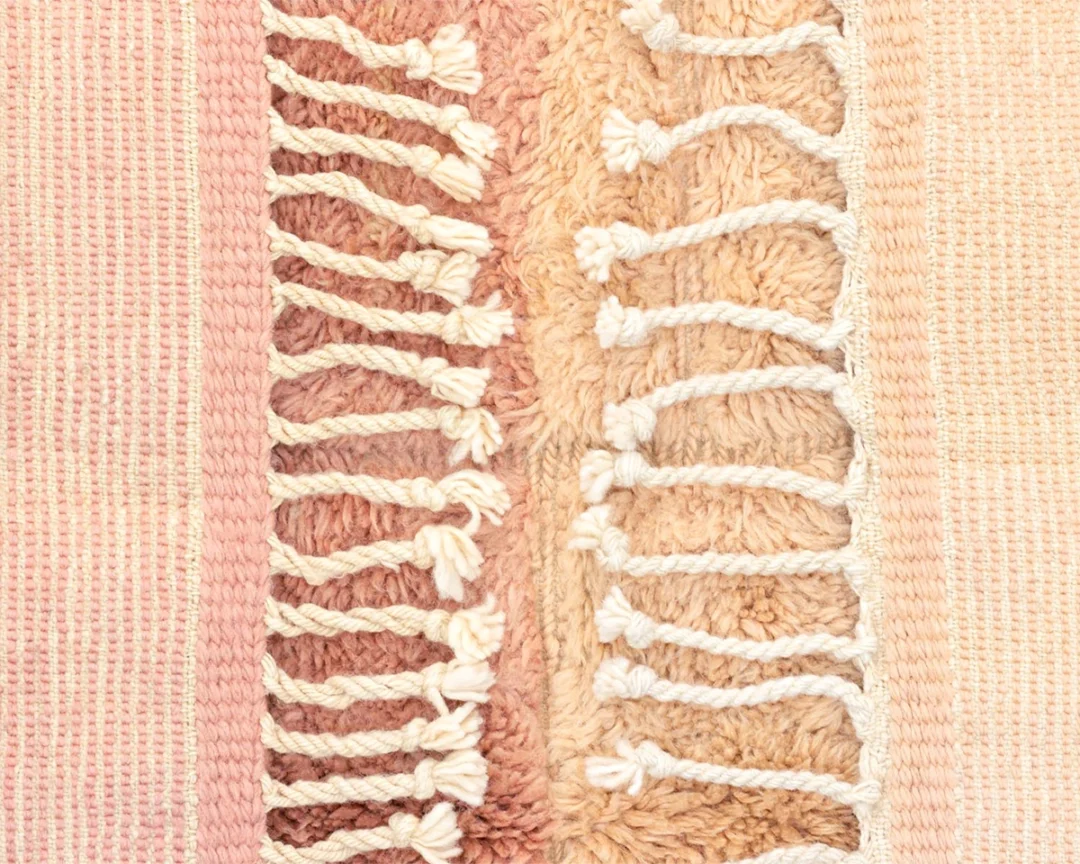 Close-up of a woven textile with pink and beige tones, featuring textured patterns and white knotted fringe running vertically down the center.