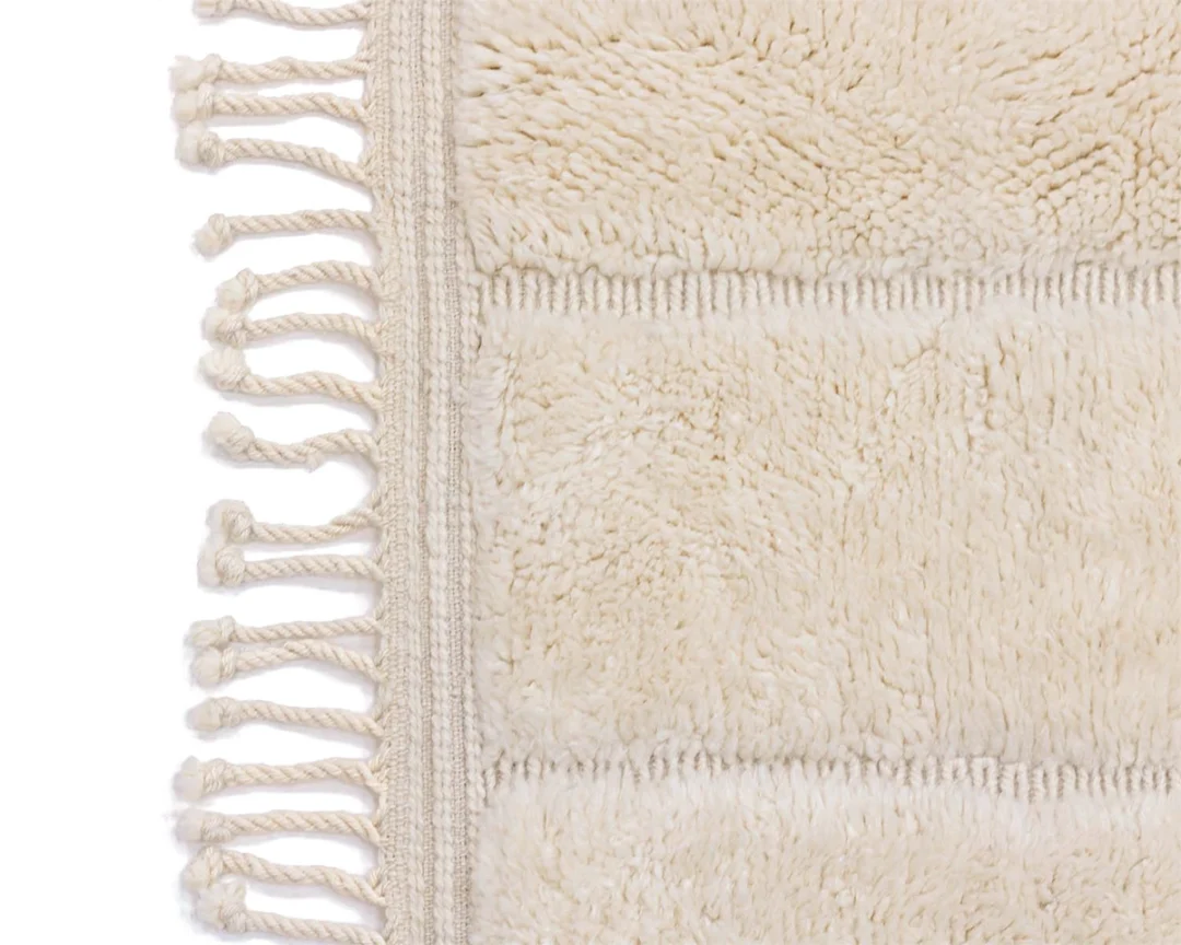 Close-up of a cream-colored, textured rug with fringed edges on the left side and a soft, plush surface on the right.