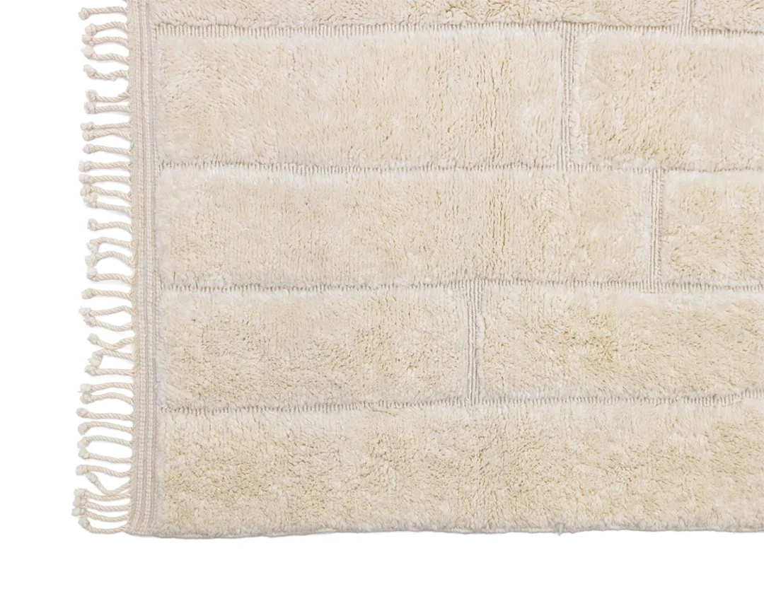 A close-up of a rectangular beige textured rug with fringe along the edge and a grid-like stitched pattern.