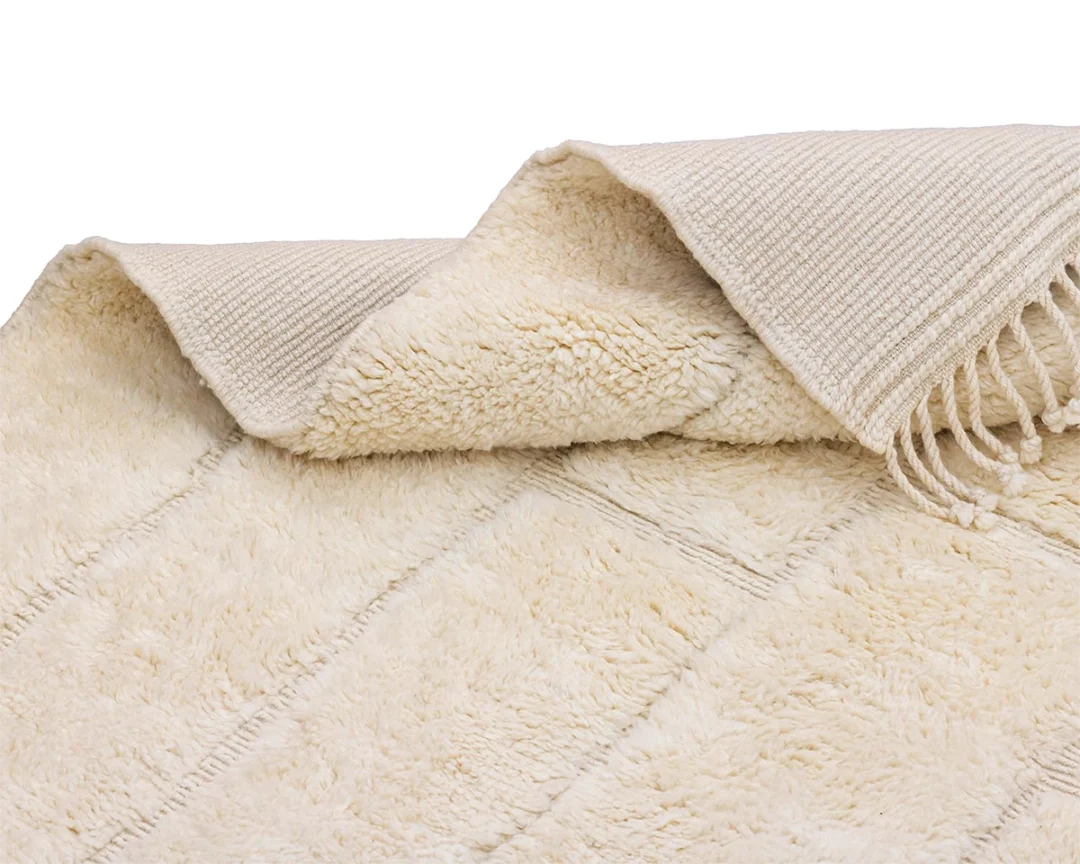 A close-up of a folded cream-colored, textured rug with fringe on one edge, showing both the soft pile and woven underside.
