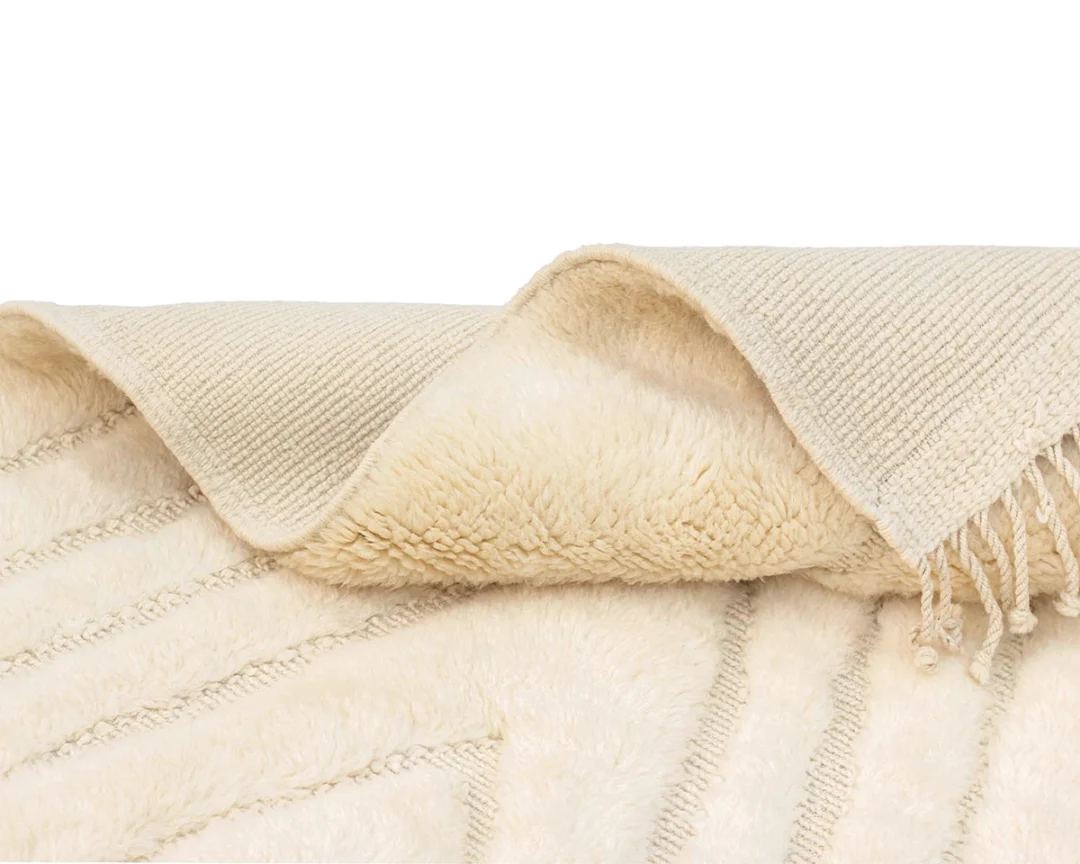 A close-up of a beige textured rug, showing both its soft, fluffy side and woven underside with fringe detail.