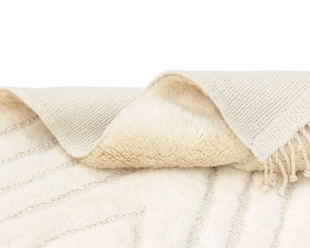A close-up of a cream-colored textured rug with a soft, plush underside and a ribbed, woven top layer with fringe on the edge.