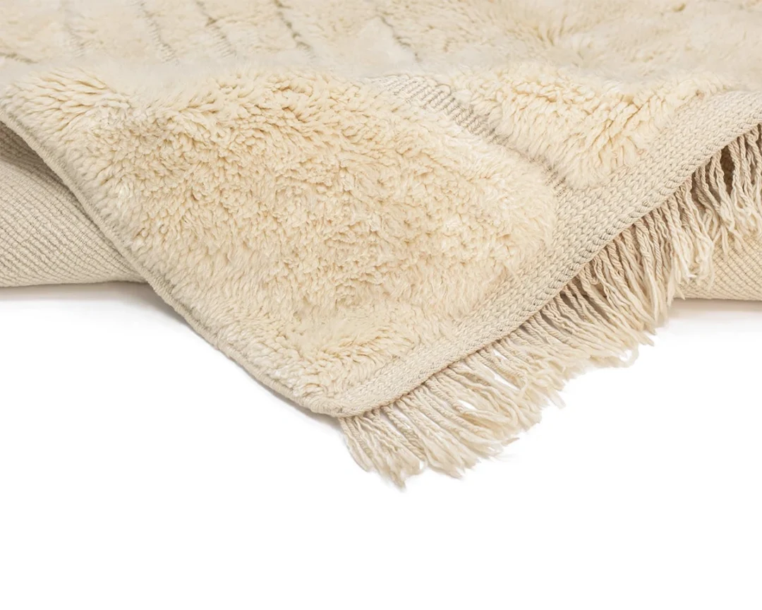 A close-up of a beige, fringed, textured throw rug with a soft, plush surface and knotted rug underside.