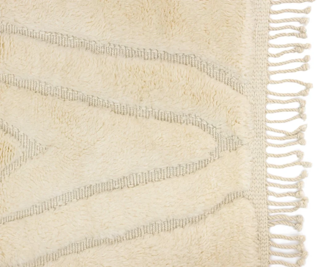 Close-up of a cream-colored textured rug with raised geometric lines and fringe tassels along the edge.