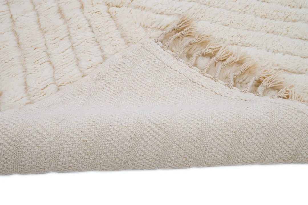 A close-up view of a cream-colored textured rug, partially folded to show the woven underside and fringed edge.