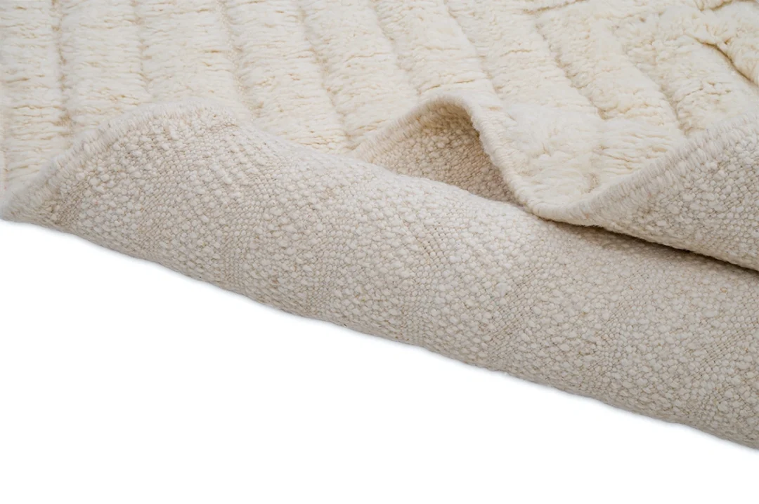 Close-up of a cream-colored, textured fabric rug with one edge folded over, showing both the top and underside patterns.
