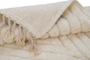 A close-up of a folded cream-colored blanket with a textured woven top layer and fringe tassels, revealing a soft, fluffy underside.