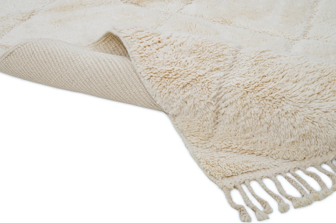 A close-up of a cream-colored textured rug with a soft, shaggy surface, visible woven underside, and fringed tassels along the edge.