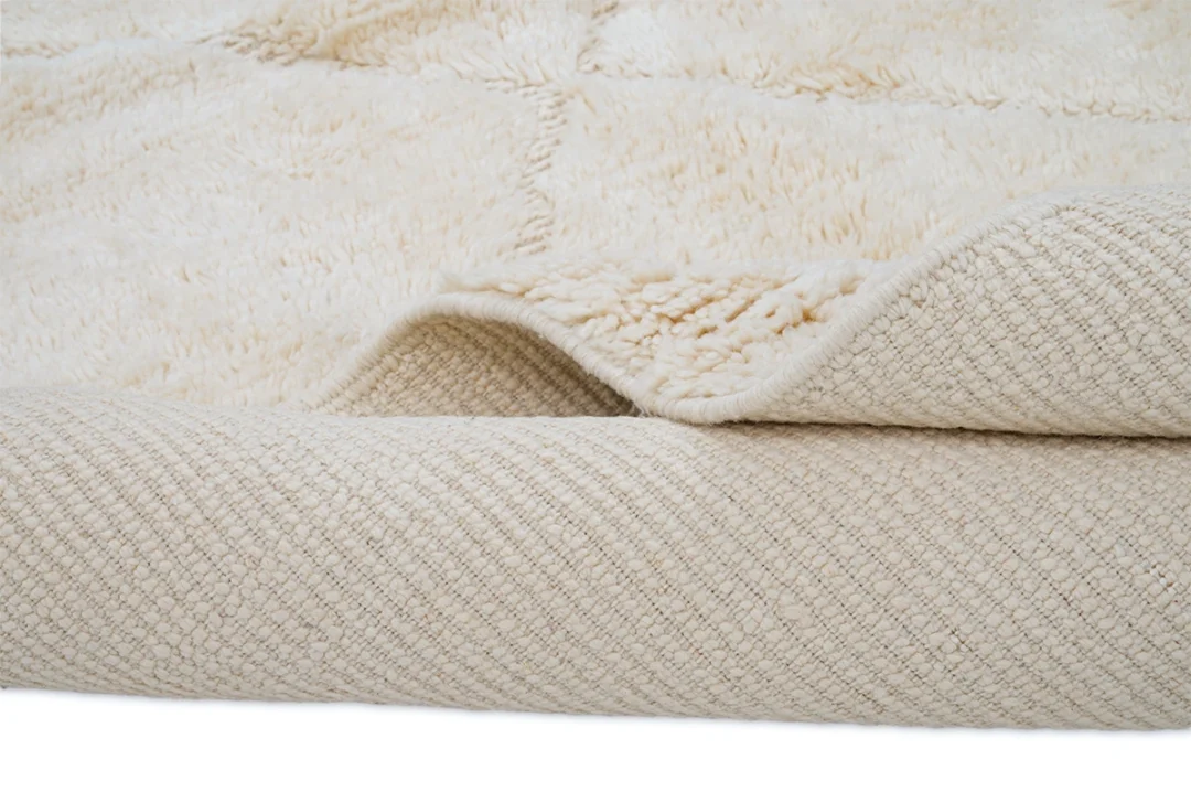 A close-up view of a cream-colored textured rug with a soft, fluffy surface and a woven underside, partially folded to show both sides.