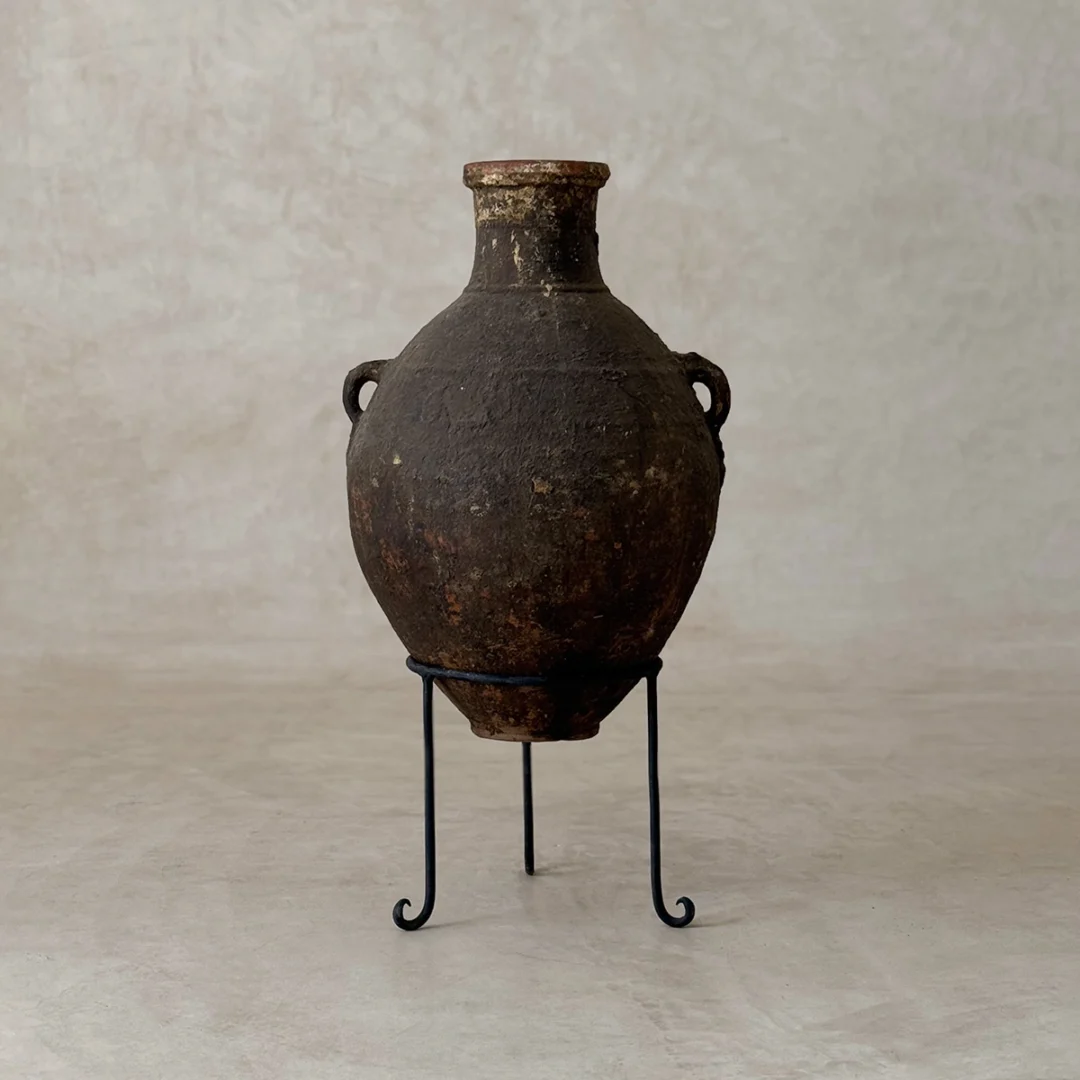 A weathered, dark brown clay amphora with two handles is displayed on a simple black metal stand against a neutral background.