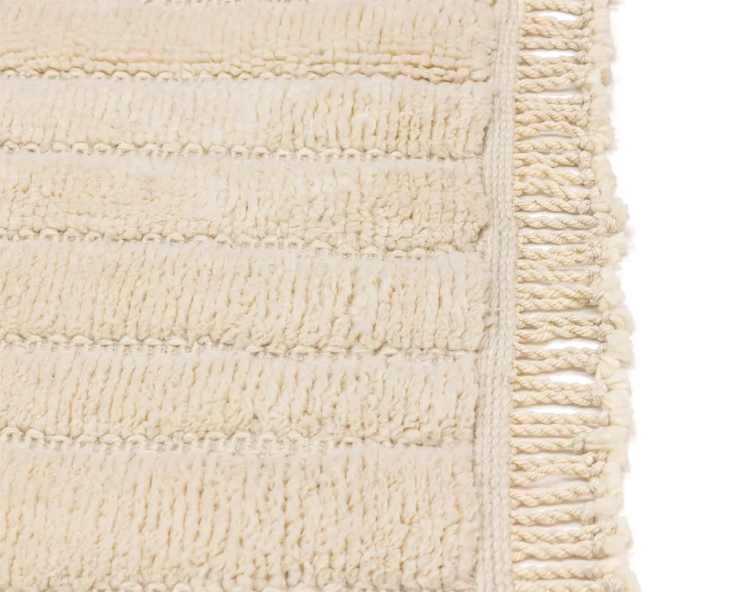 Close-up of a cream-colored textured fabric rug with horizontal lines and fringe detailing on one edge.