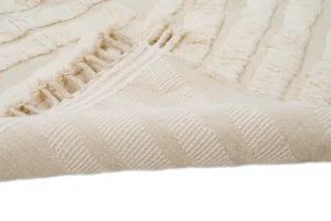 A close-up of a beige textured rug with a folded corner, showing its woven underside and tasseled edge.
