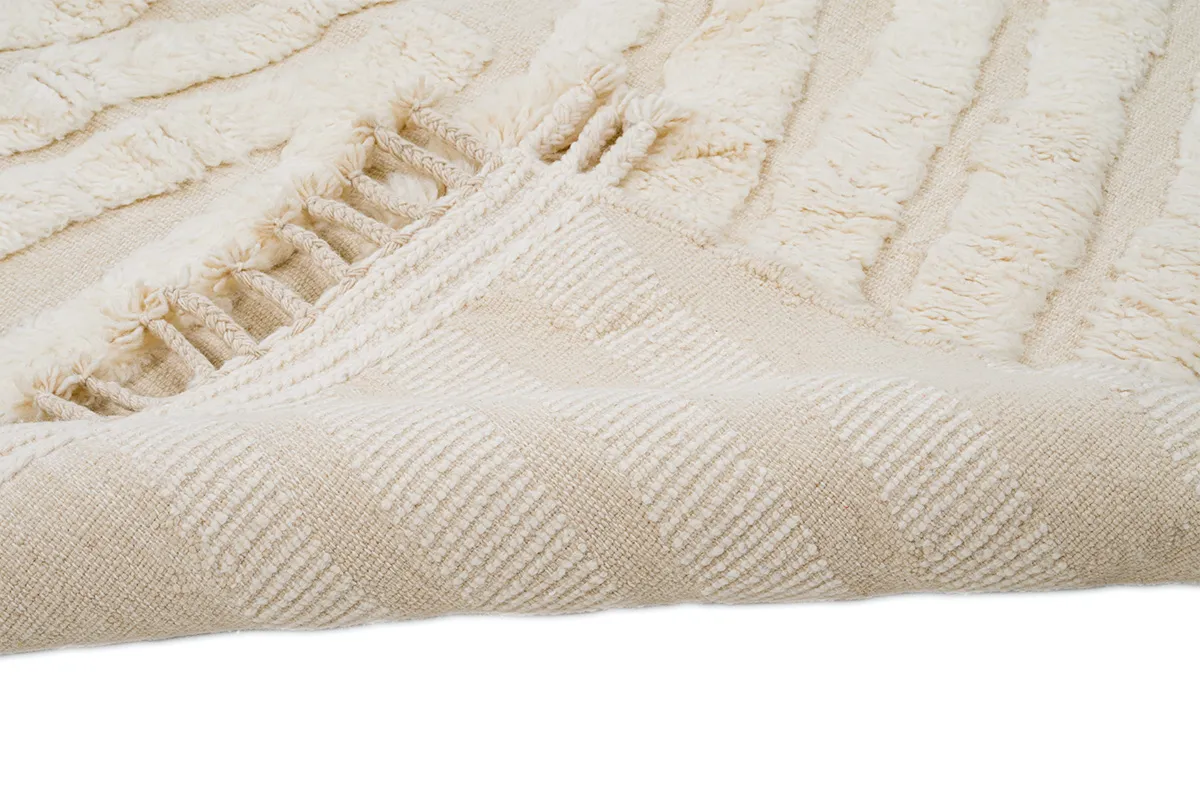 A close-up of a beige textured rug with a folded corner, showing its woven underside and tasseled edge.