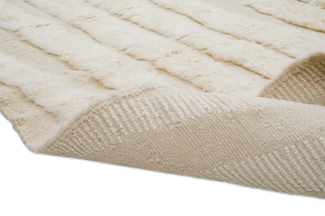 A close-up of a cream-colored textured rug with raised, fluffy stripes and woven detailing, showing the corner turned up.