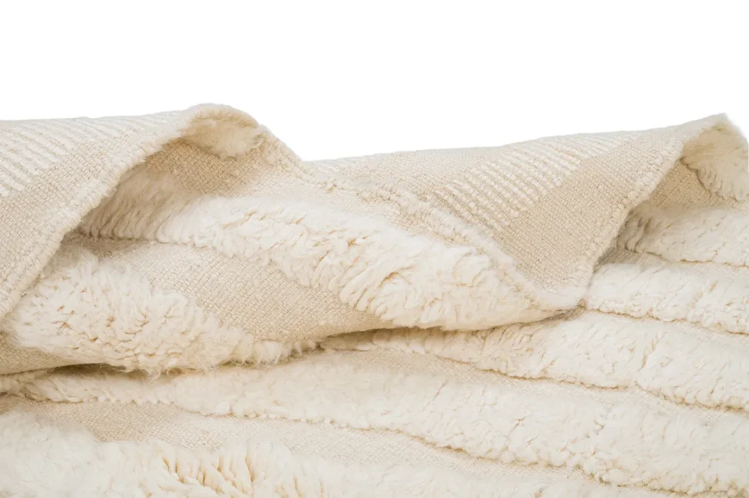 A close-up of a beige textured rug with soft, fluffy and ribbed rug folds against a white background.