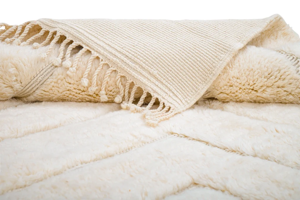 A close-up of a cream-colored textured blanket with a ribbed woven edge and tassels, laid out on a matching plush surface.