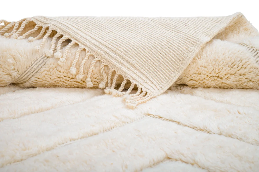 A close-up of a cream-colored textured blanket with a ribbed woven edge and tassels, laid out on a matching plush surface.
