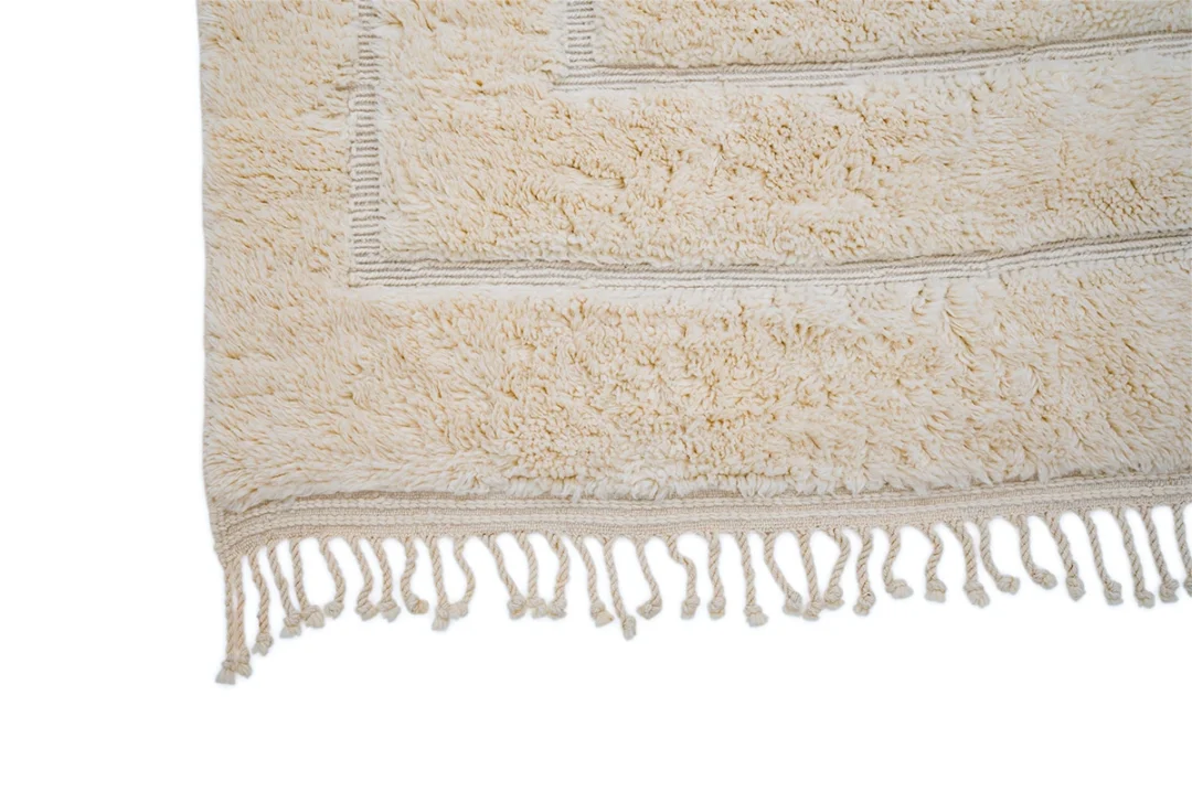 Close-up of the corner of a beige, textured rug with a fringed edge on a white background.