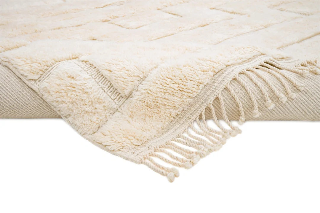 A close-up of a beige, textured rug with raised patterns and fringed edges shown against a white background.