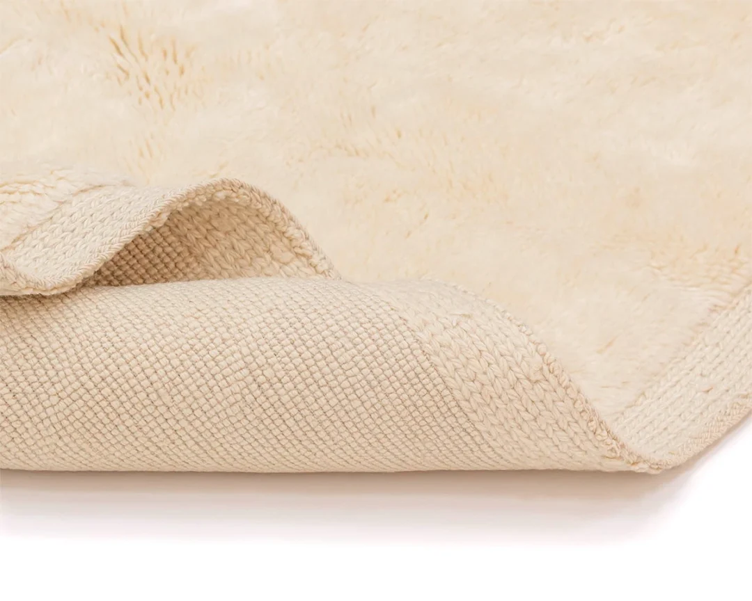 A close-up view of a beige rug partially folded back, showing its textured backing and soft, plush surface.