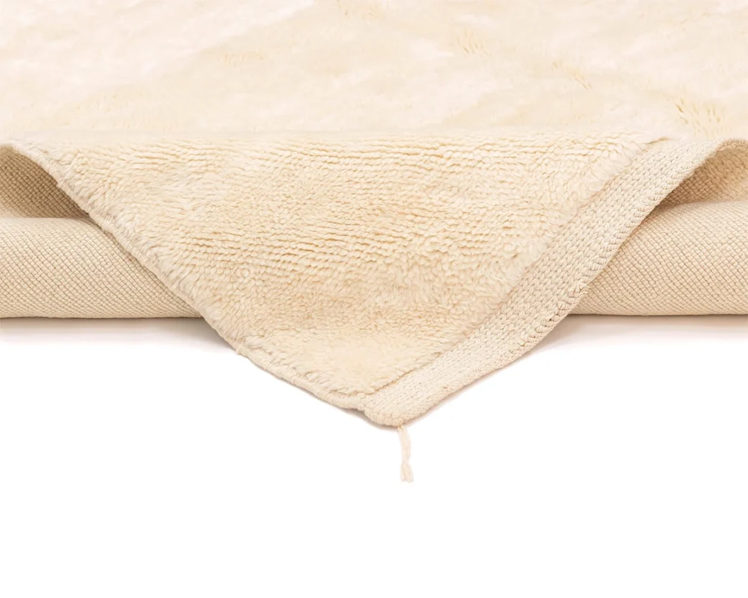 A folded beige rug with a textured surface and a visible corner draped forward, showing both sides of the rug against a white background.