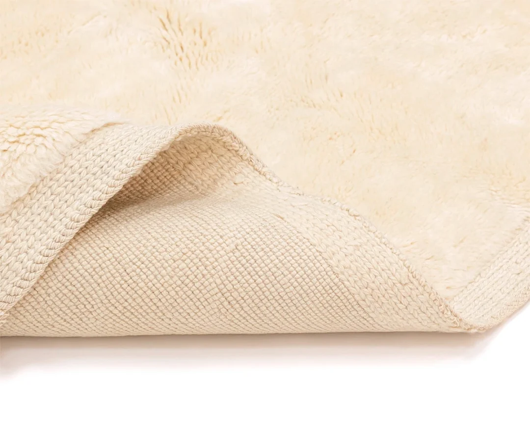 A close-up of a cream-colored, textured rug with one corner flipped over, showing both the soft top surface and the woven underside.