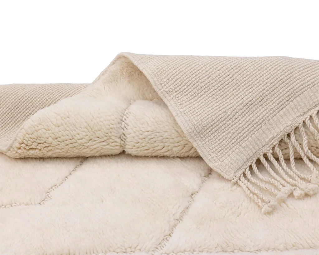 A folded cream-colored textured throw blanket with a soft, plush underside and fringed edges is shown on a white background.